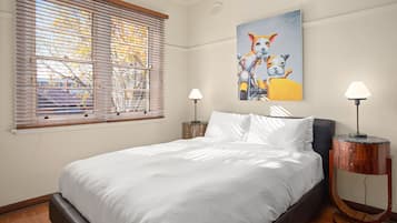 A bedroom with a bed, two bedside lamps, a window with blinds, and a painting on the wall.