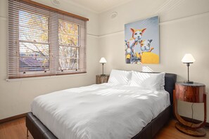 A bedroom with a bed, two bedside lamps, a window with blinds, and a painting on the wall. - Cherrybombe 1920s Art Deco Armadale Apartment (Armadale)