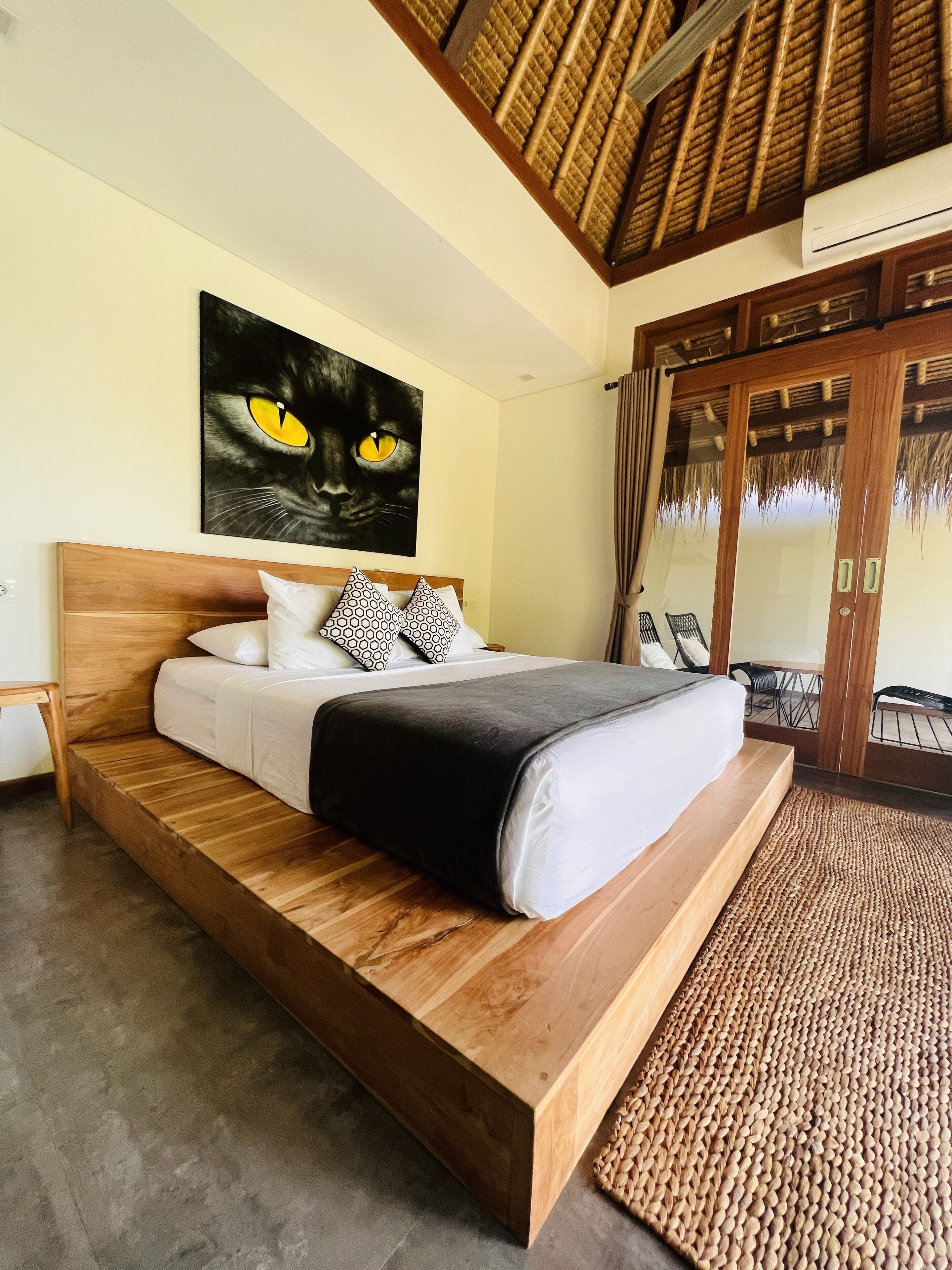 A modern bedroom with a wooden bed, a large painting of a cat's face, and a view of the outside through a glass door.
