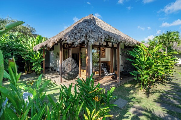 A thatched-roof structure with a glass door, surrounded by lush greenery.