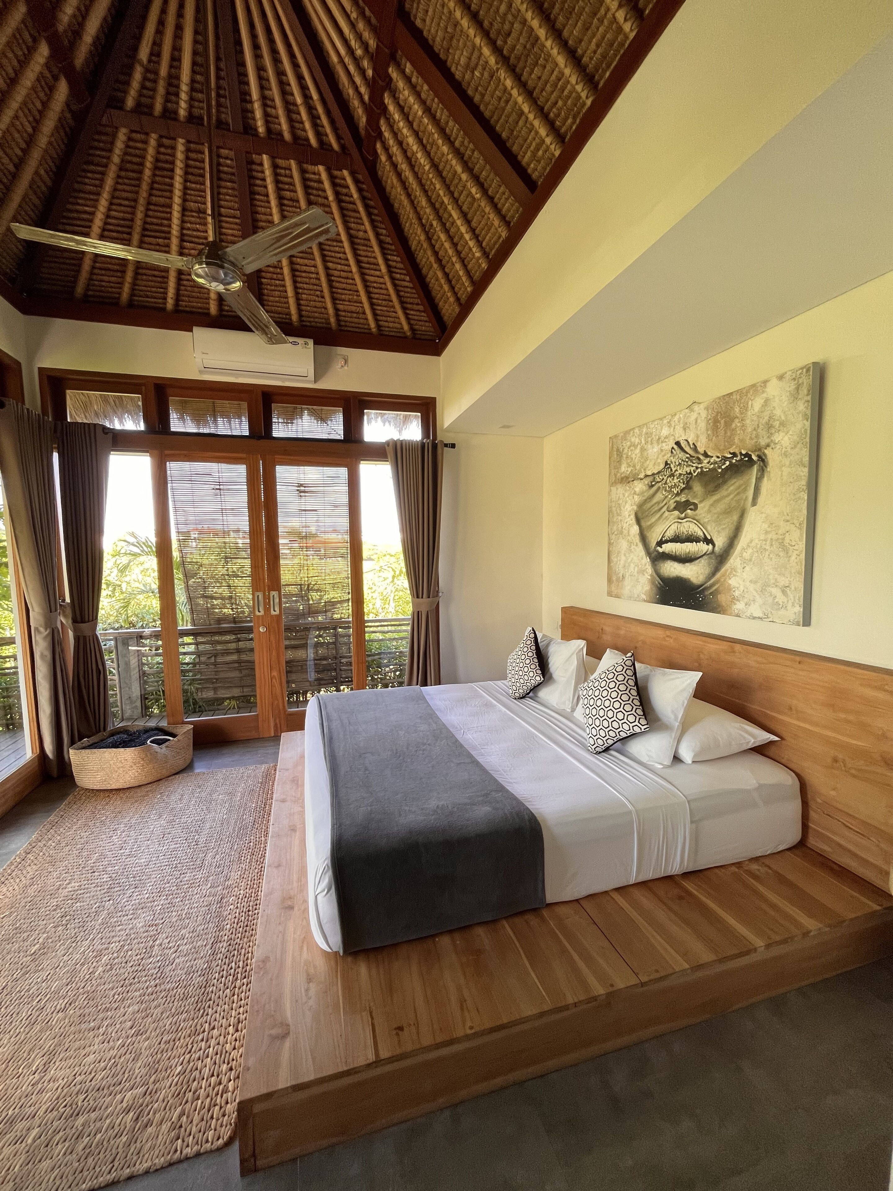 A bedroom with a bed, a ceiling fan, a large window with curtains, and a painting on the wall.