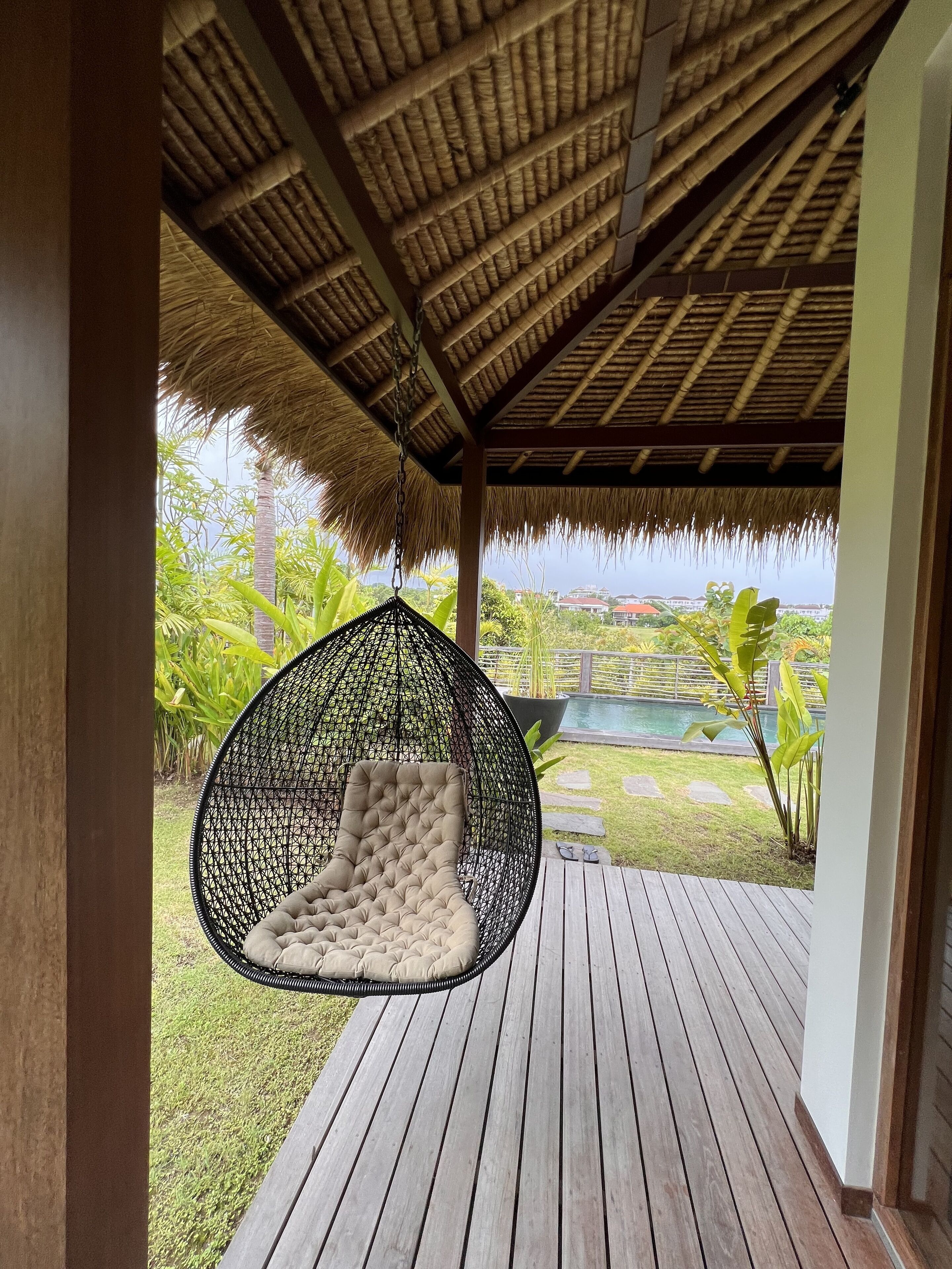 Un espacio al aire libre con techo de paja, un sillón colgante y vistas a una piscina y follaje.