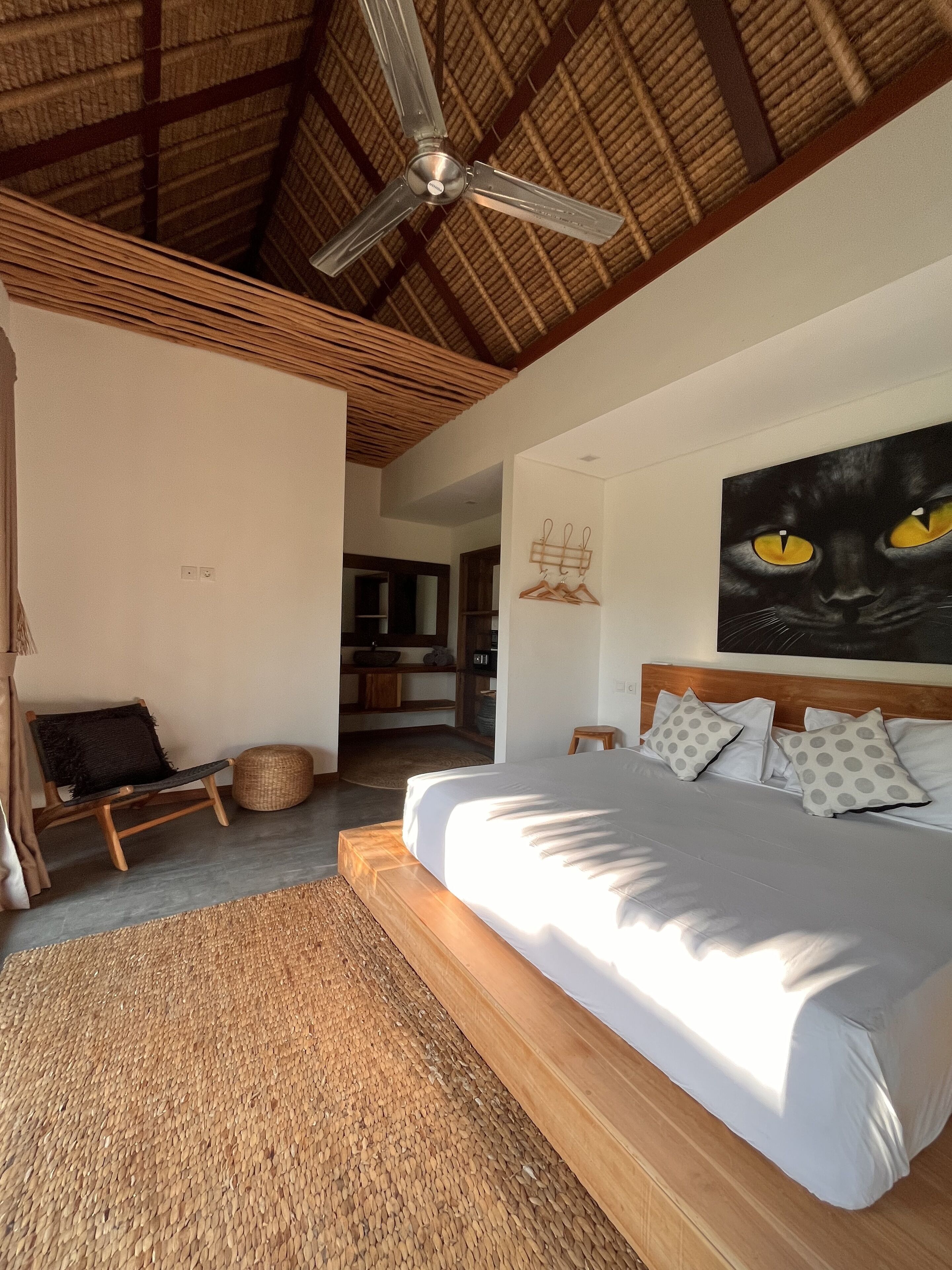 A bedroom with a bed, a ceiling fan, a painting of a cat, and a view into another room.