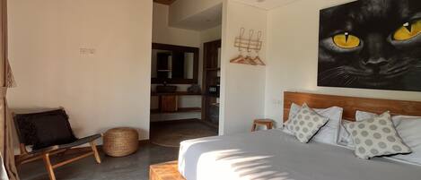 A bedroom with a bed, a ceiling fan, a painting of a cat, and a view into another room.