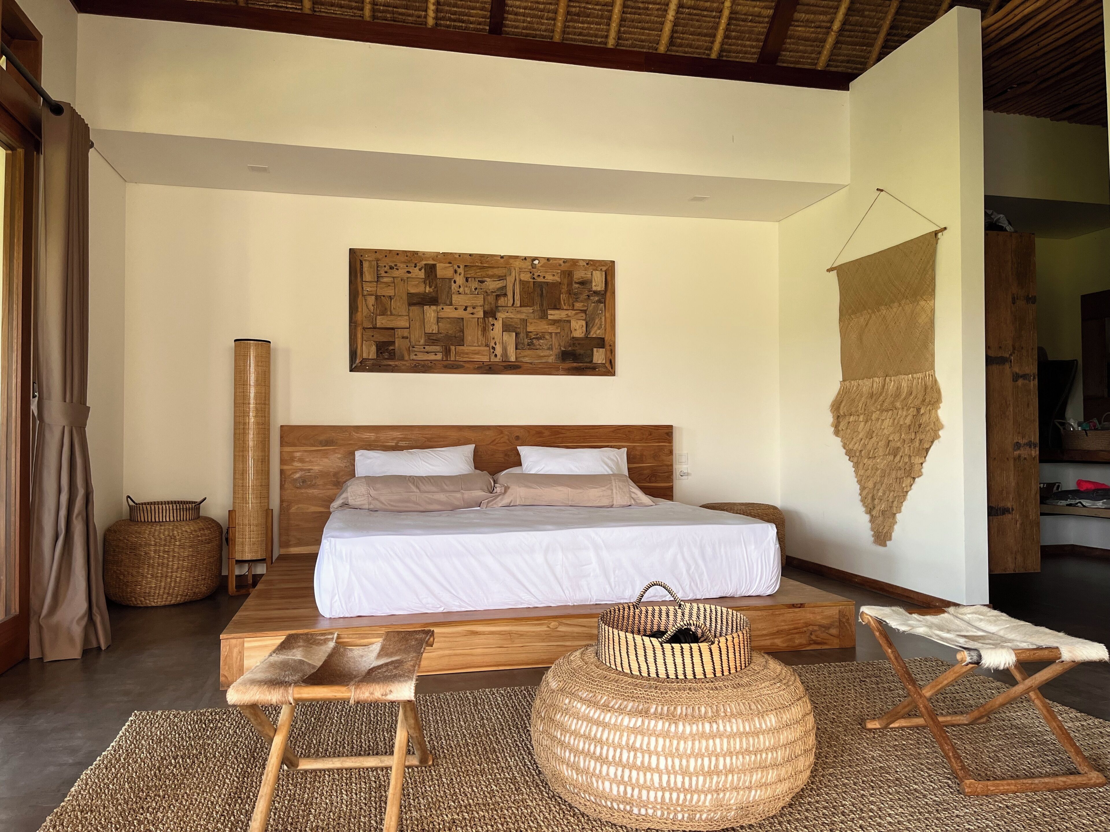 A bedroom with a wooden bed, a woven headboard, a woven pouf, a wooden bench, and a woven pouf on the floor.