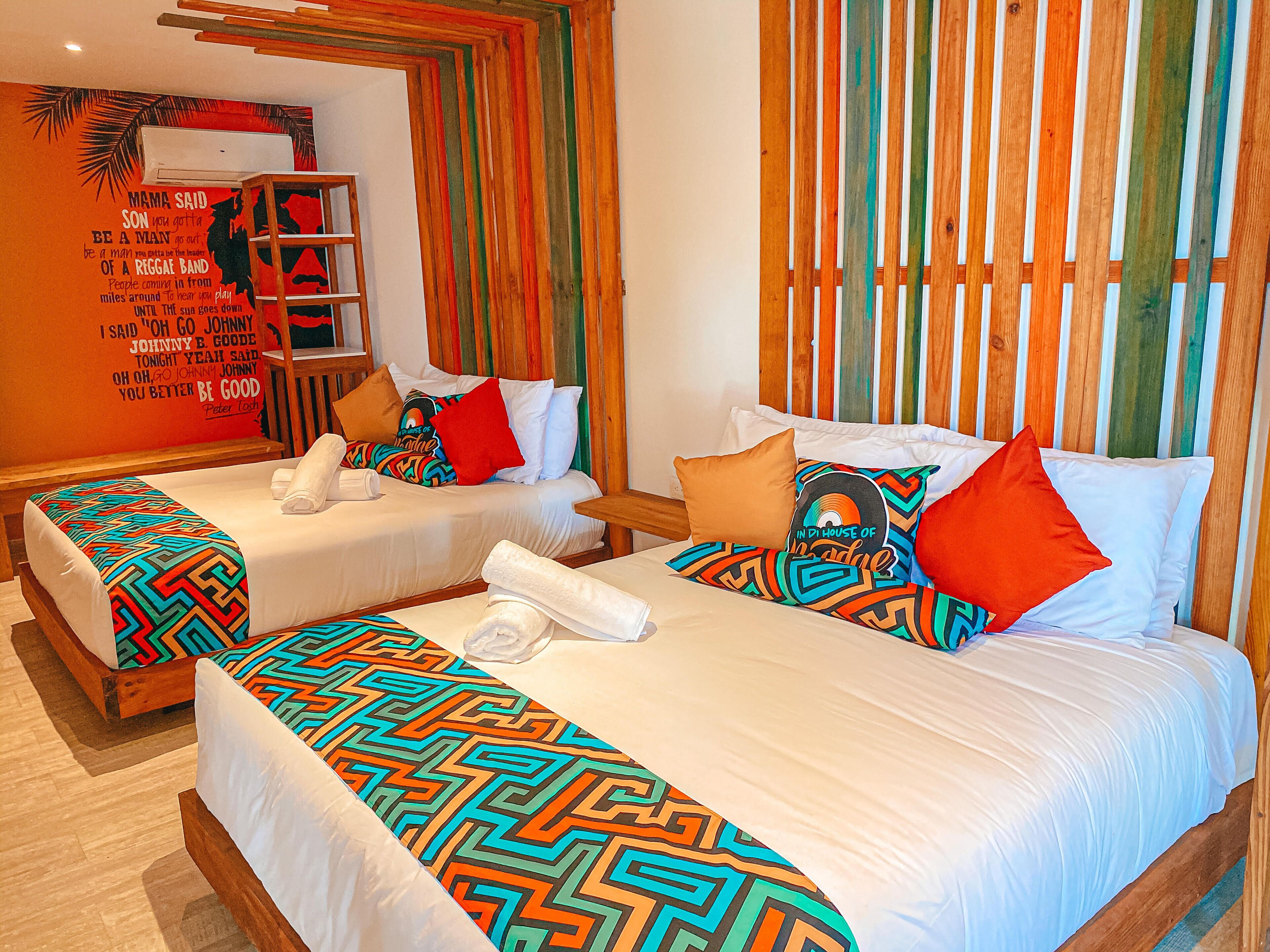 A hotel room with two beds, each with patterned bedspreads and pillows, a wooden headboard, a wall with a quote, and a shelf with towels.