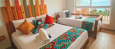 A double bed with a colorful geometric bedspread, a wooden headboard, and a window with a view of the ocean.
