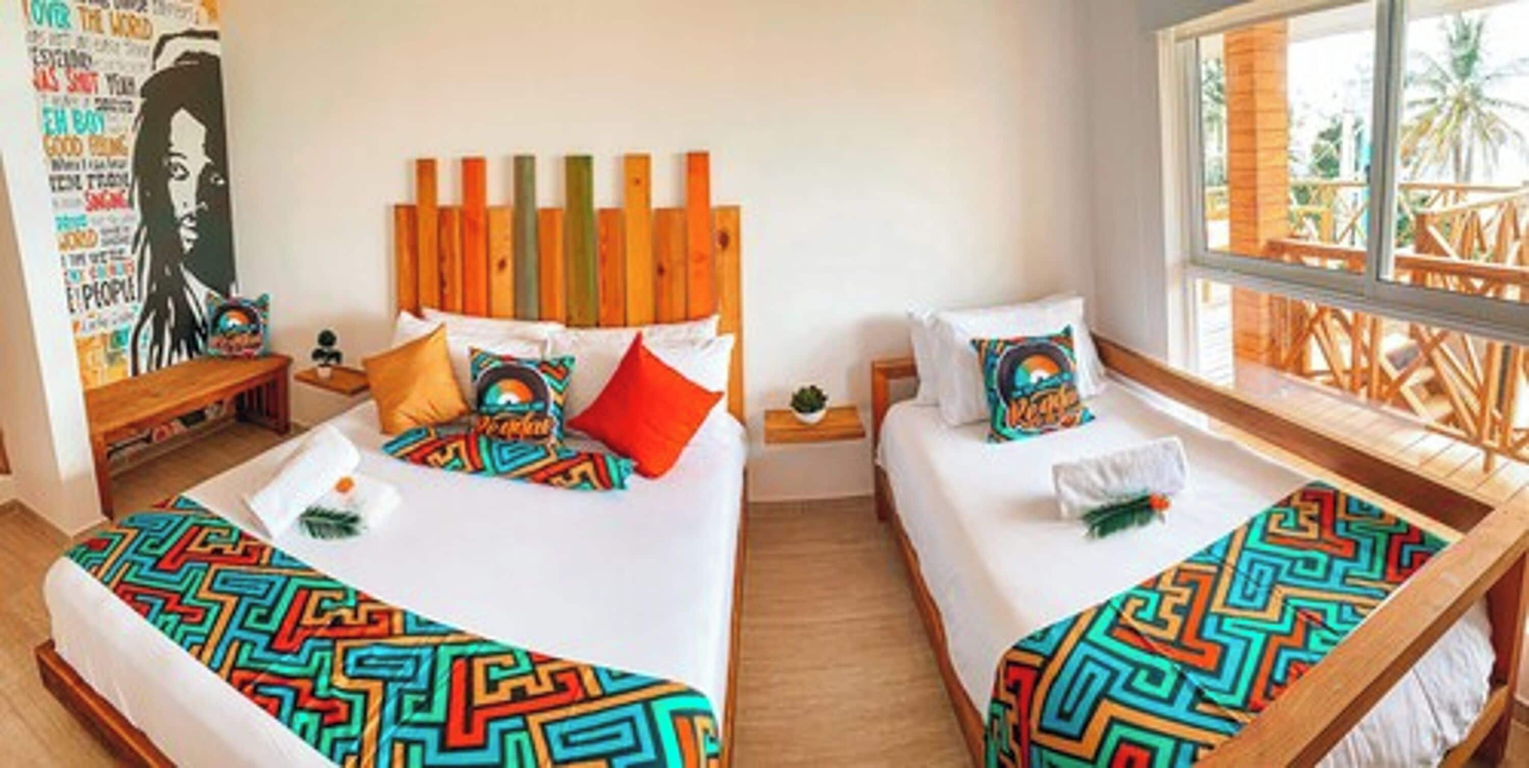 A hotel room with two beds, colorful patterned bedspreads, a wooden headboard, a wall with a poster, and a window with a view of palm trees.