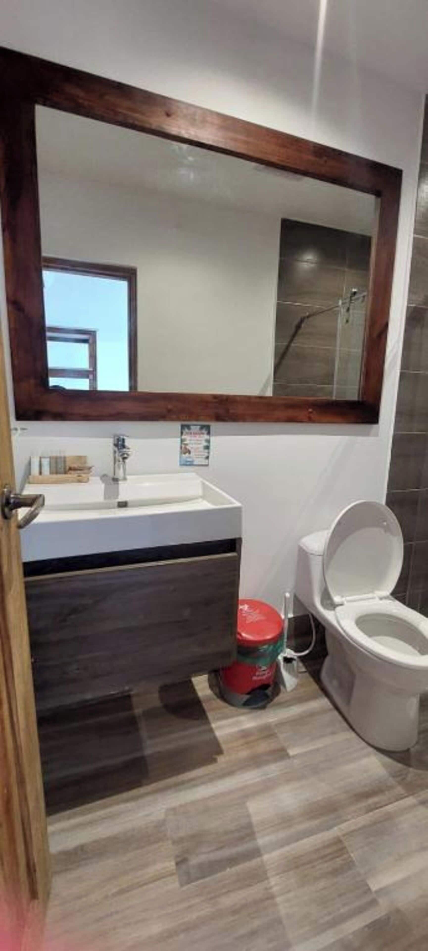 A bathroom with a wooden-framed mirror, a white sink, a dark wooden cabinet, a toilet, and a red bucket.