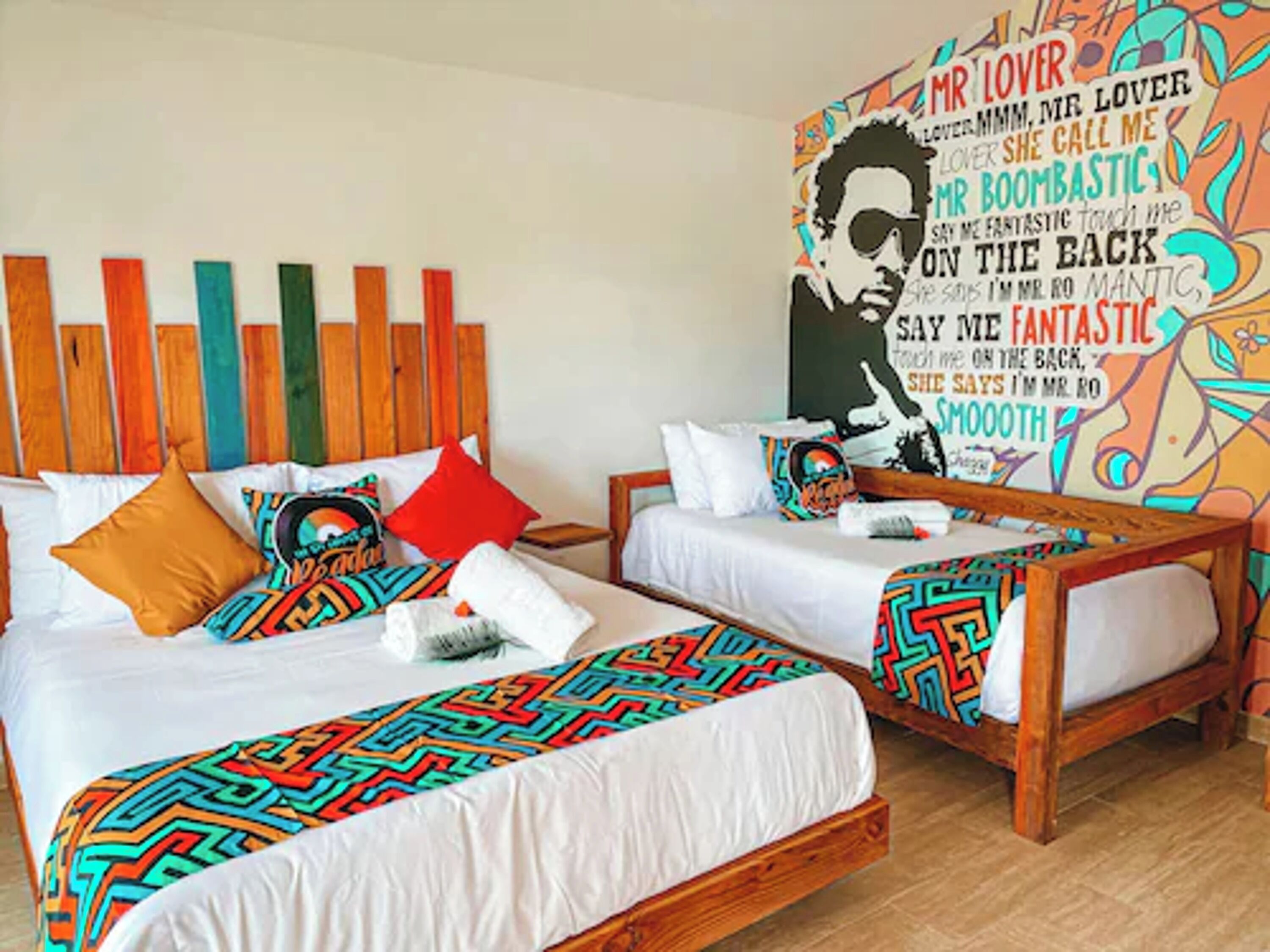 A hotel room with two beds, colorful bedding, a wooden headboard, and a vibrant wall mural.