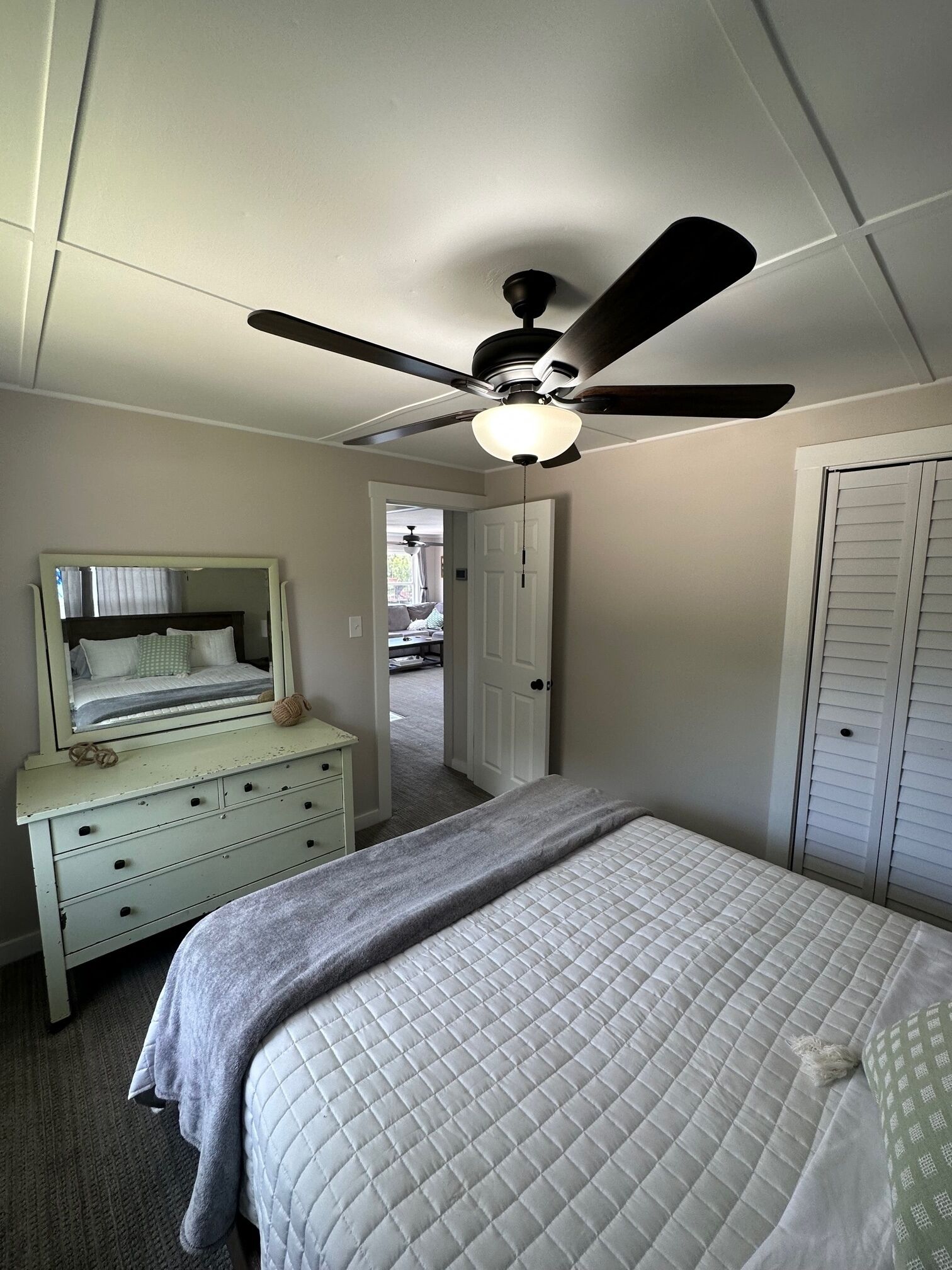 A bedroom with a bed, a dresser, a ceiling fan, and a door leading to another room.