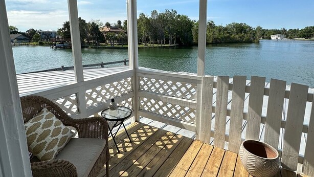 A covered outdoor deck with wicker furniture and a small table, overlooking a lake with buildings in the distance.