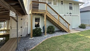 A two-story house with a wooden staircase leading to a balcony, surrounded by a gravel driveway and a grassy lawn.