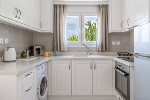 A modern kitchen with white cabinets, a washing machine, a coffee maker, and a window with a view of a cactus.
