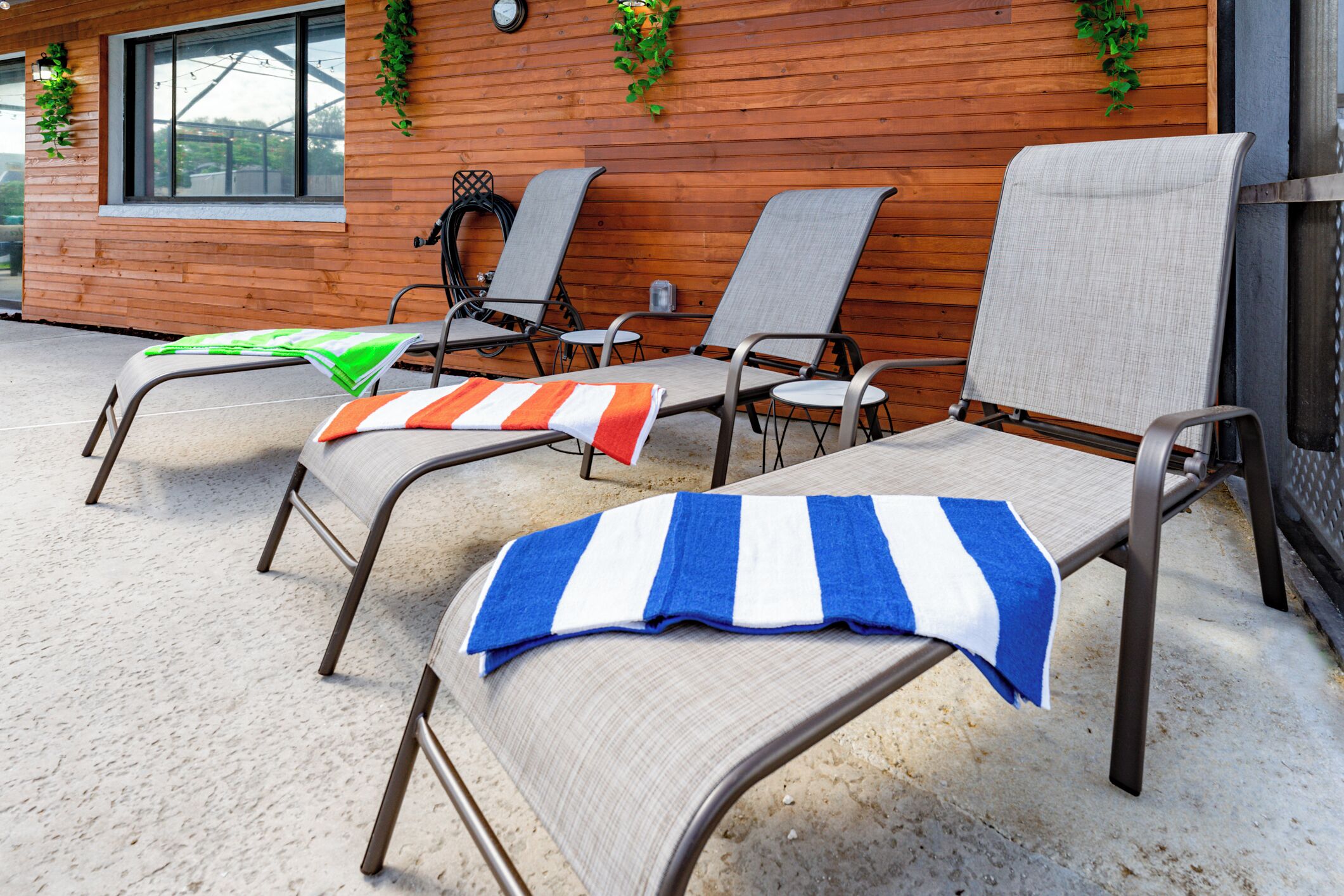 A row of sun loungers with striped towels, a wooden wall, and a window with a clock.
