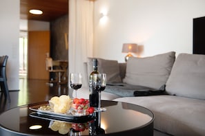 A dining table with a tray of snacks, two glasses of red wine, and a bottle of wine in a modern living room with a sofa and a lamp in the background.