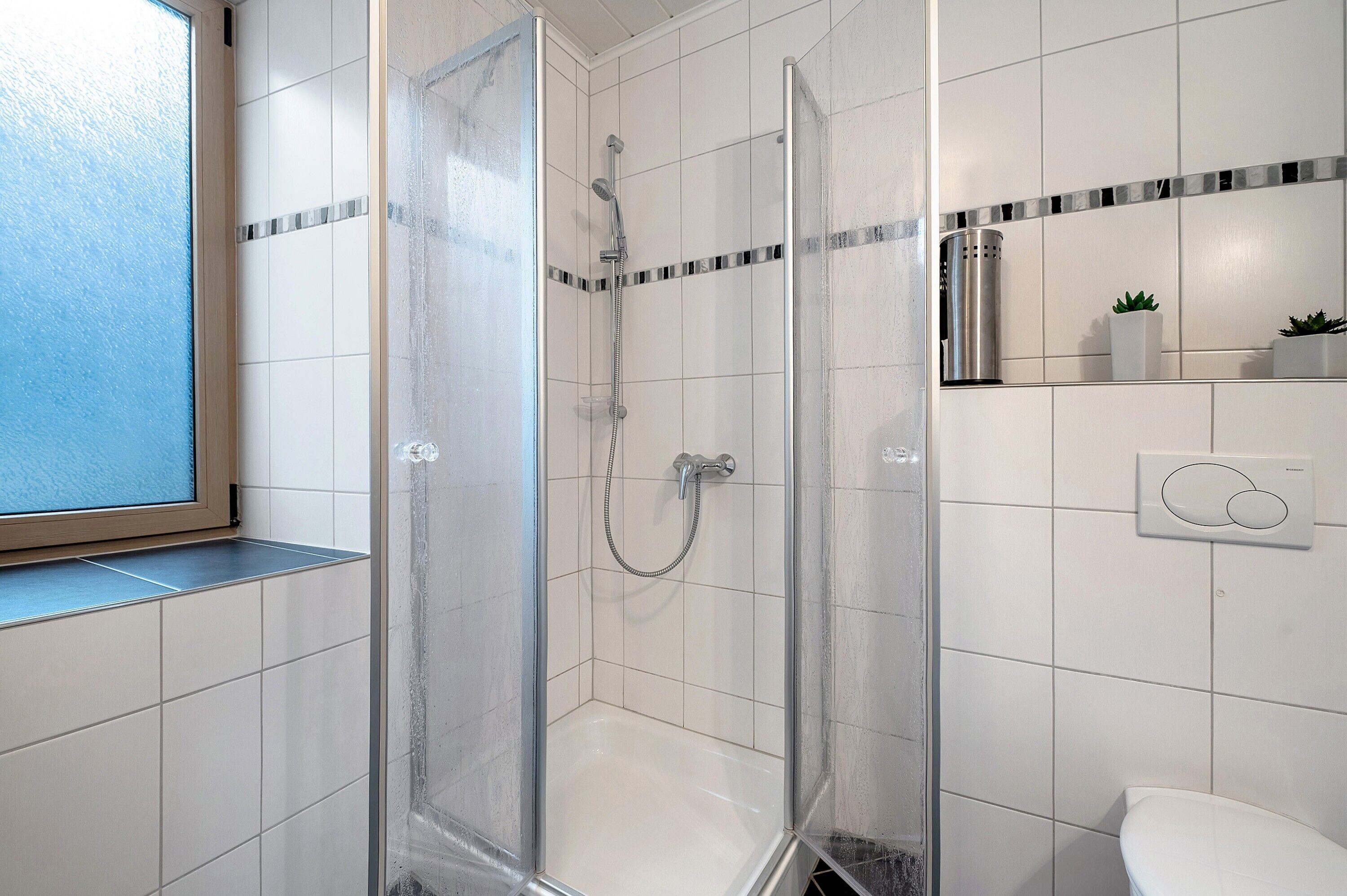 Een moderne badkamer met een douchecabine van glas, witte betegelde wanden, een toilet en een klein plankje met planten.