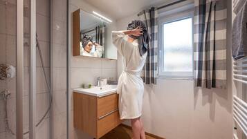 Ein Badezimmer mit Duschkabine aus Glas, einem Holzbadezimmermöbel, einem Spiegel und einem Fenster mit Vorhängen.