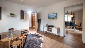 A modern living room with a wooden dining table, a sofa, and a large rug. There is a TV, a shelf with plants, and a painting on the wall. - Apartment 'Eve' with Mountain View, Balcony and Wi-Fi (Söll)