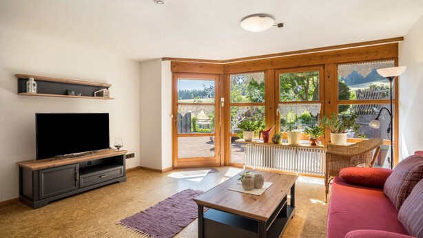 A living room with a TV, a coffee table, a red sofa, and large windows with a view of greenery and mountains.