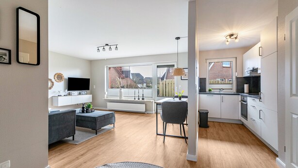 A modern kitchen with white cabinets, a dining area with a grey sofa, and a large window offering a view of the outside. - Apartment "Haus Dünenbutze" with Shared Garden & Wi-Fi (Büsum)