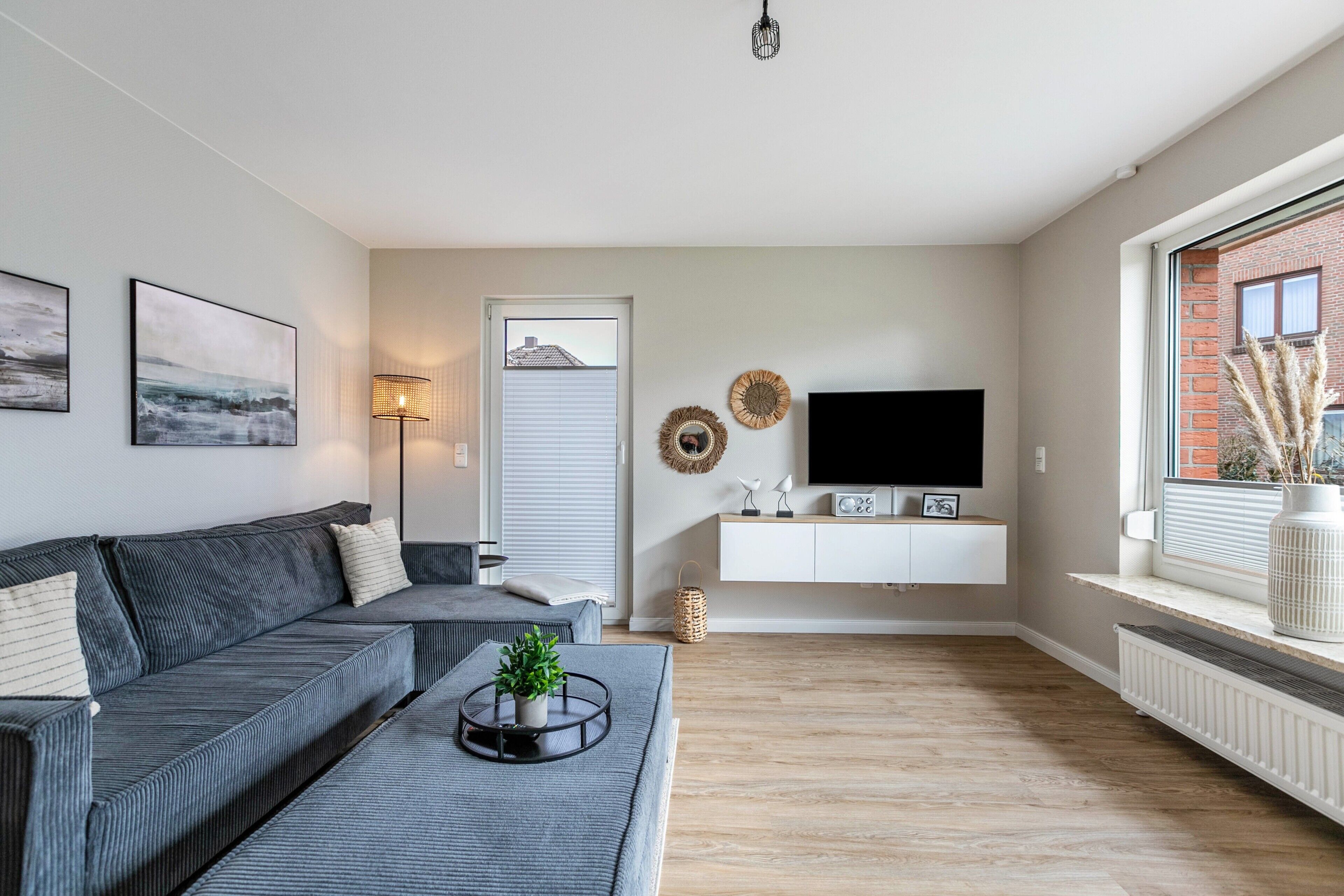 Een moderne woonkamer met een grijze bank, een bijzettafeltje met een plant, een flatscreen-tv op een wit kastje en een raam met uitzicht op een bakstenen gebouw.