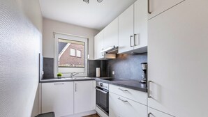 A compact kitchen with white cabinets, a black countertop, a window with a view of a brick building, and a small potted plant. - Apartment "Haus Dünenbutze" with Shared Garden & Wi-Fi (Büsum)