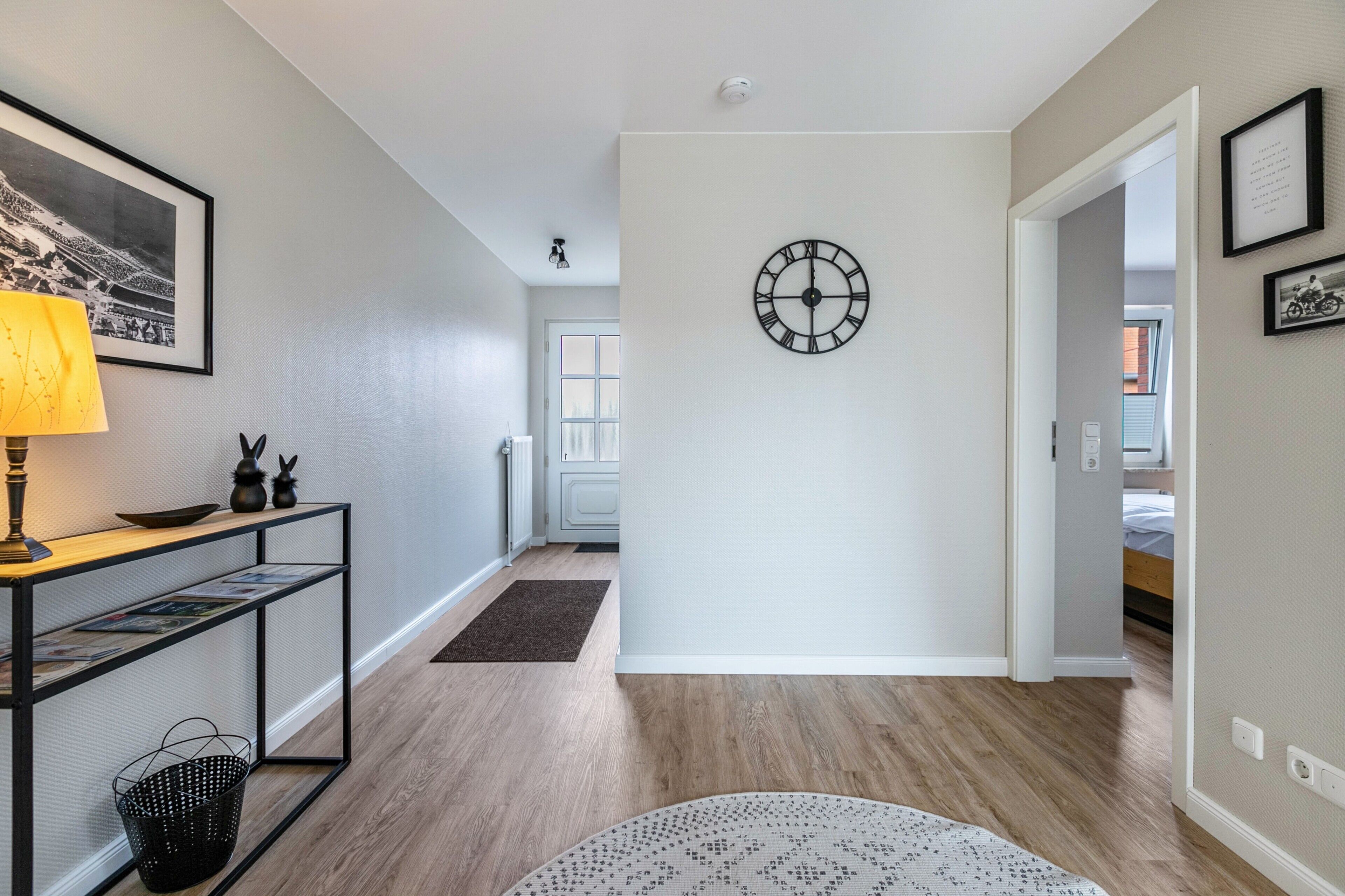 Un couloir avec un plancher en bois, une horloge murale, un tableau encadré et une lampe posée sur une console.