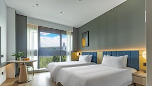 Quarto de hotel moderno com duas camas, uma mesinha com uma planta, vista da cidade e uma janela grande com cortinas.
