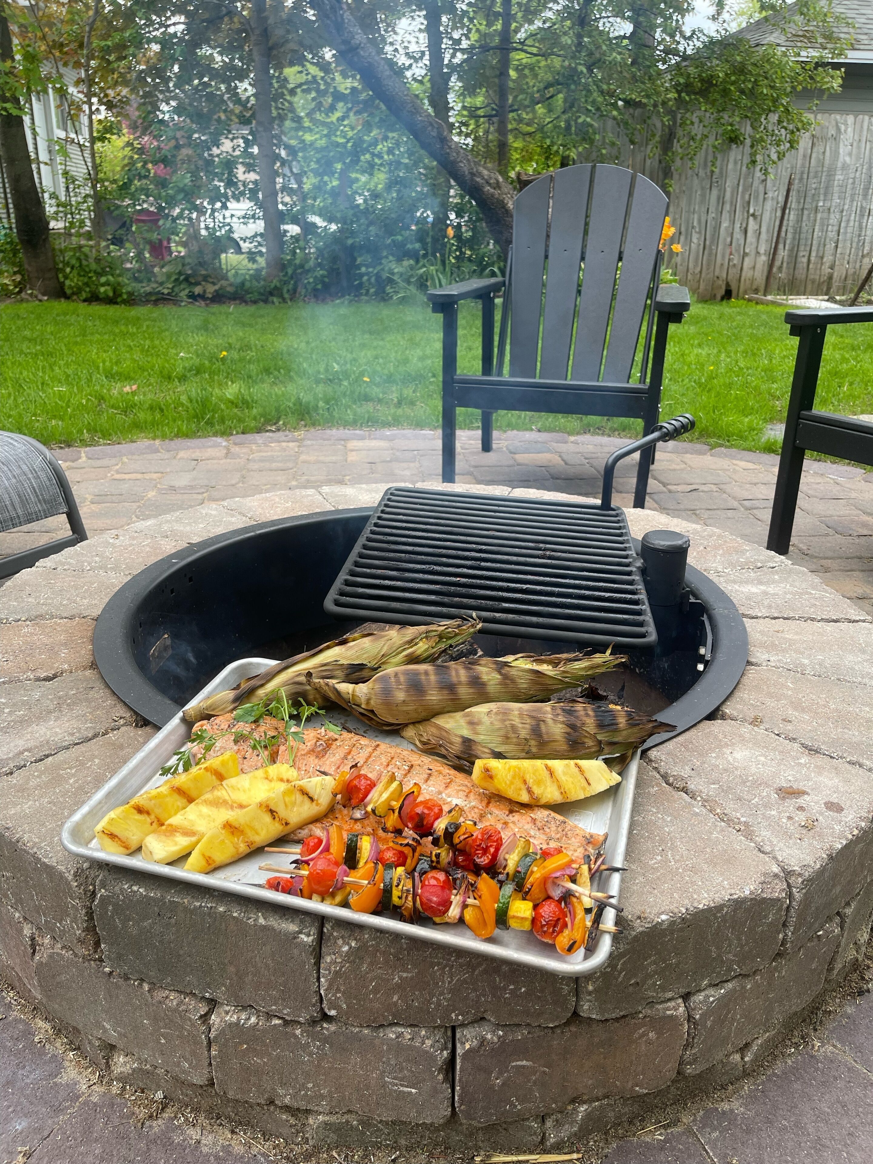 Een buitenbarbecue met een vuurkorf van natuursteen, een metalen dienblad met gegrilde groenten en maïs, en twee Adirondack-stoelen op de achtergrond.