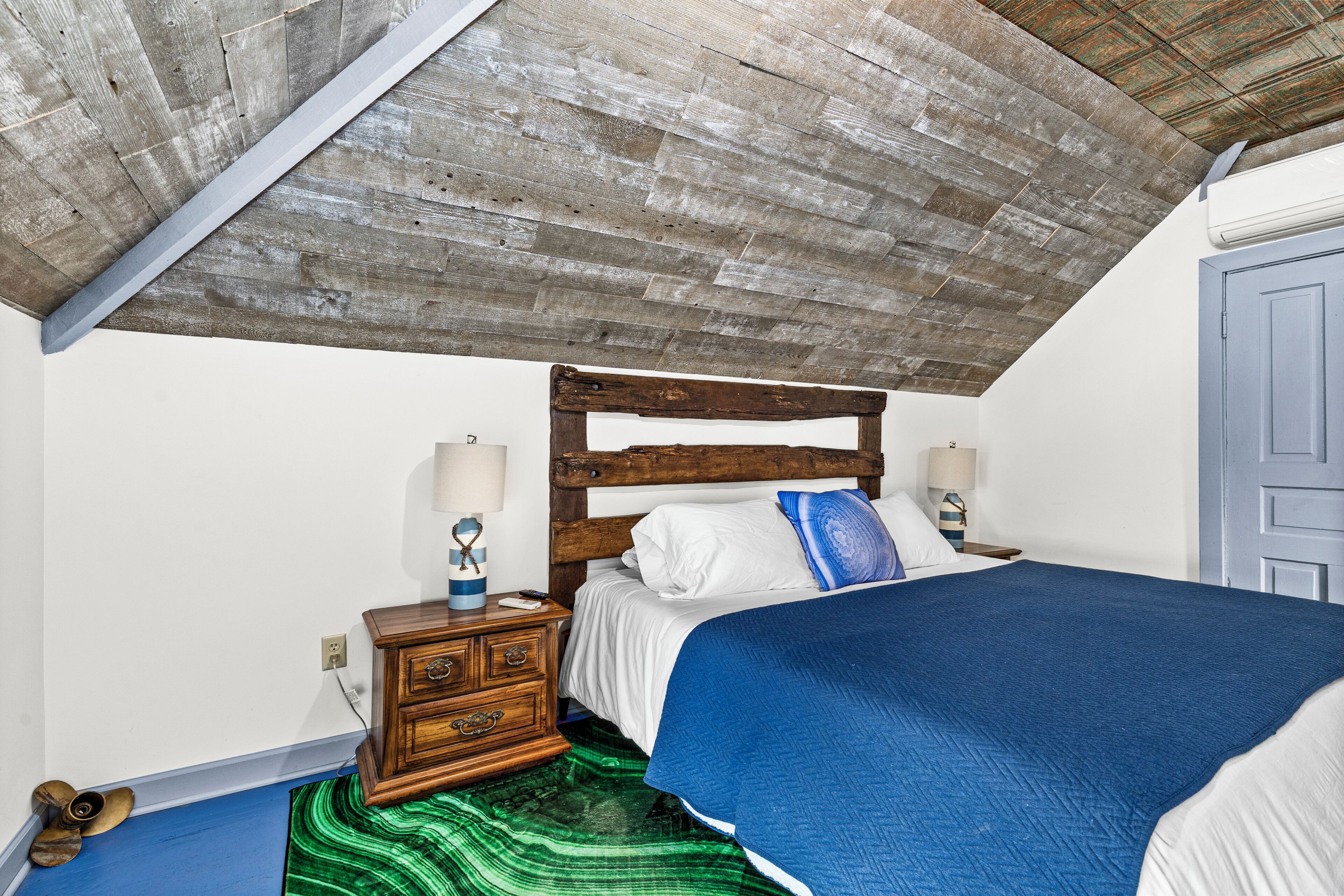 Ein Schlafzimmer mit Holzboden, einem Bett mit blauer Decke, einem Nachttisch mit Lampe und einem grün gemusterten Teppich.