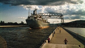 Un barco grande pasando por debajo de un puente sobre una masa de agua, con gente en el muelle.