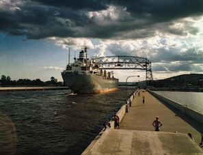 Ein großes Schiff passiert eine Brücke über dem Wasser, während sich Menschen an der Kaimauer befinden.