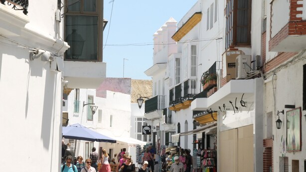 A narrow street with white buildings, some shops, and pedestrians. - Acuario Apartment in Conil (Conil)
