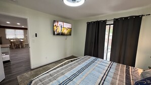 A bedroom with a bed, a TV mounted on the wall, a ceiling fan, and a view of the outside through a window with curtains.