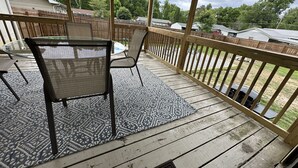 A covered outdoor deck with a table and chairs, a patterned rug, and a wooden railing.
