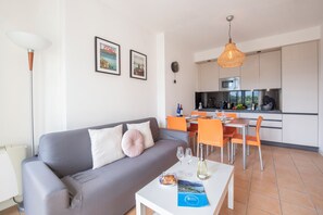 Una sala de estar moderna con un sofá gris, una mesa de comedor con sillas naranjas, una cocina al fondo y cuadros en la pared.