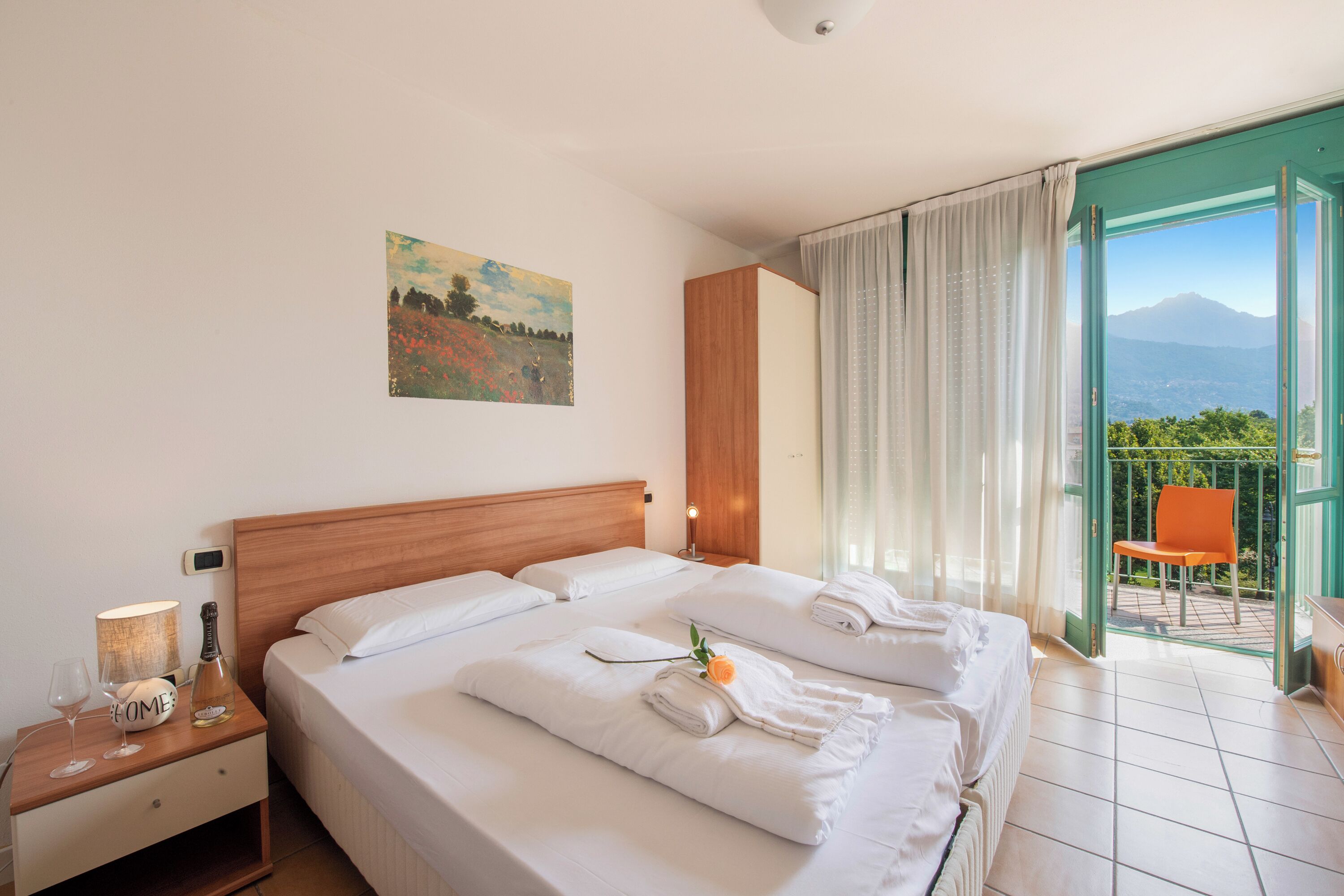 Une chambre d’hôtel avec un grand lit, une table de chevet avec une lampe et une bouteille, un tableau au mur et un balcon avec une chaise.