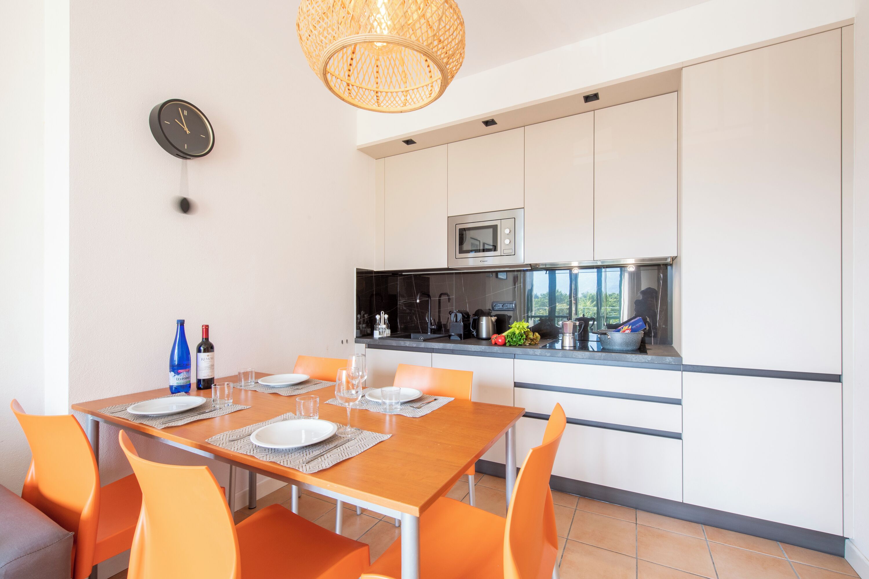 A modern kitchen with white cabinets, a microwave, and a sink. There is a dining table with orange chairs and a clock on the wall.