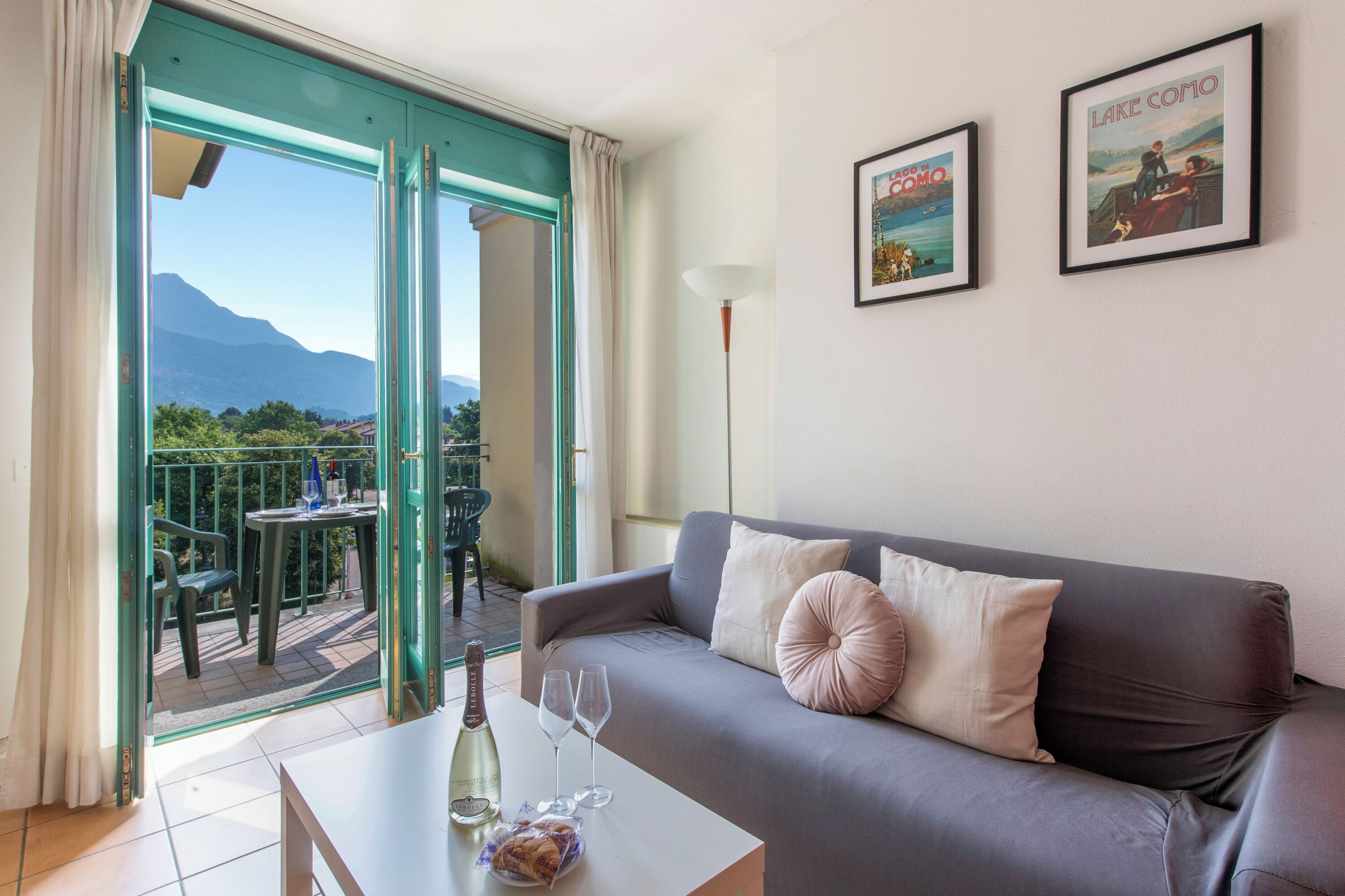 A modern living room with a grey sofa, two cushions, a table with a bottle and glasses, and a balcony with a table and chairs.