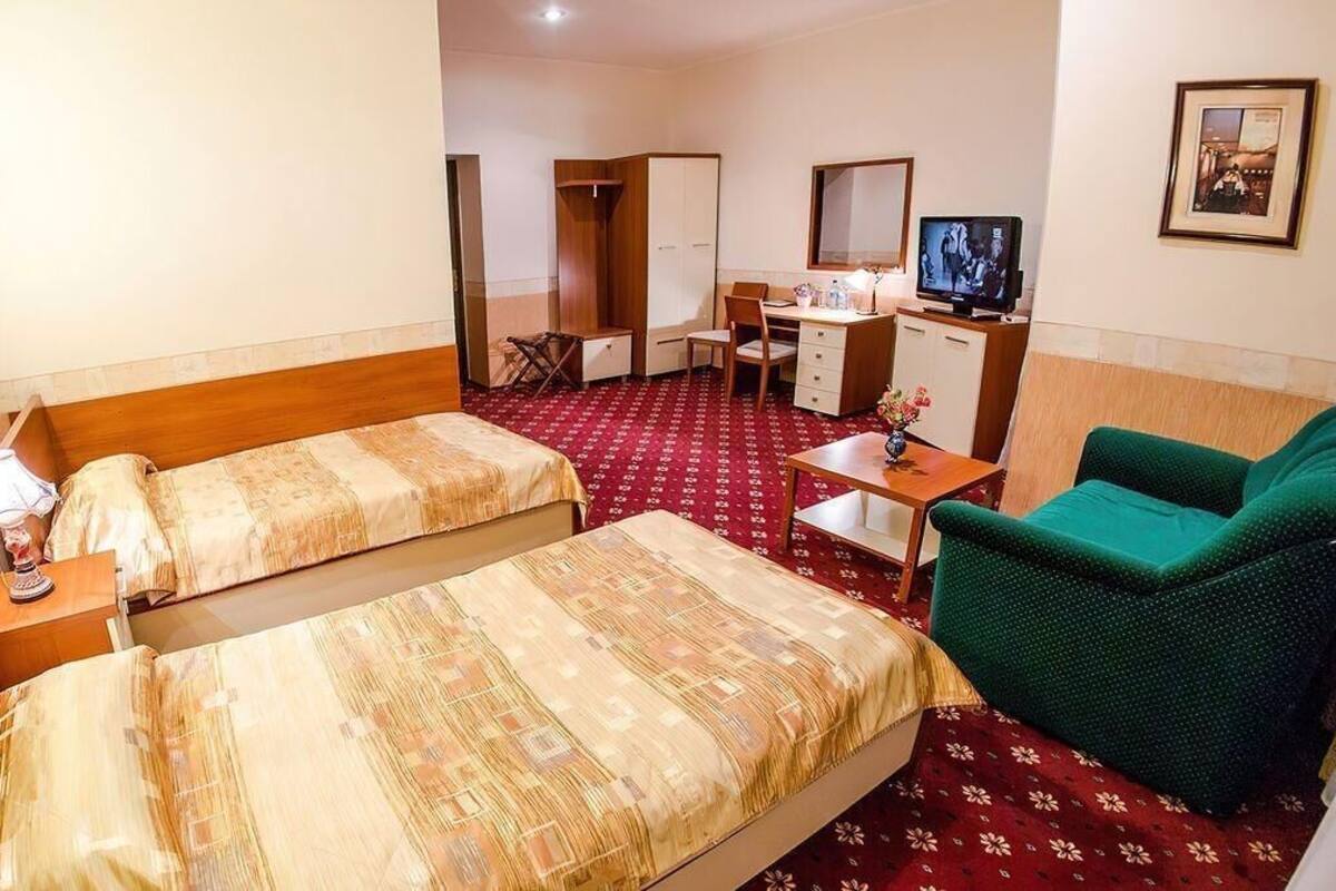 Quarto de hotel com duas camas, uma escrivaninha, uma cadeira, uma televisão e um sofá verde.