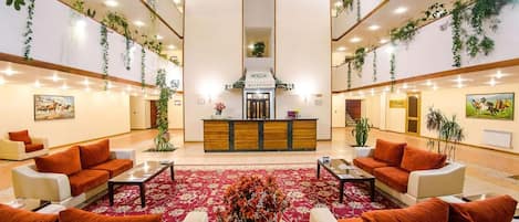 A hotel lobby with a reception desk, sofas, a red carpet, and hanging plants.