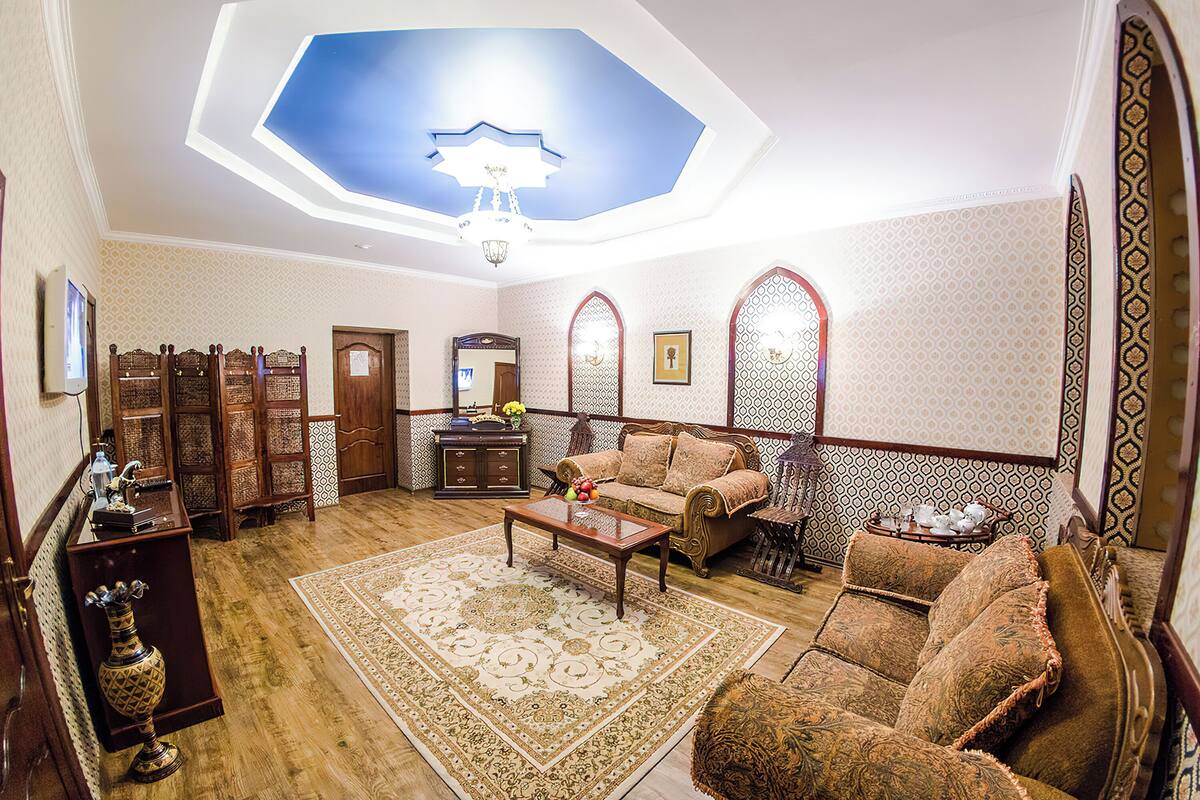 Uma sala de estar ampla com teto alto, um lustre, um conjunto de sofás, uma mesa de centro e um tapete estampado.