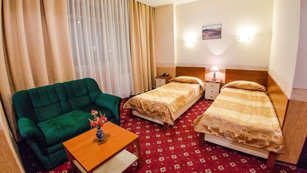 A hotel room with a green sofa, two single beds, a wooden nightstand with a lamp, a small table with a vase, and a red patterned carpet.