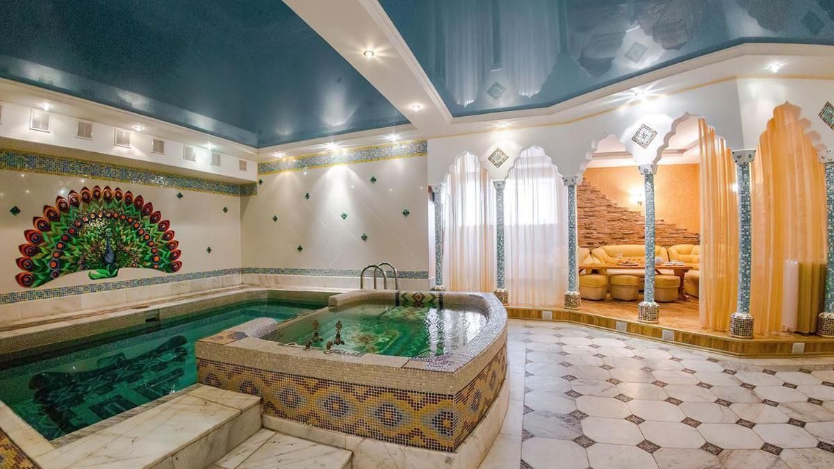 A luxurious indoor pool area with a mosaic wall, a hot tub, and a seating area with curtains.