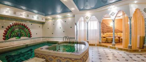 Un área de piscina interior lujosa con un muro de mosaicos, una bañera de hidromasaje y un espacio de descanso con cortinas.