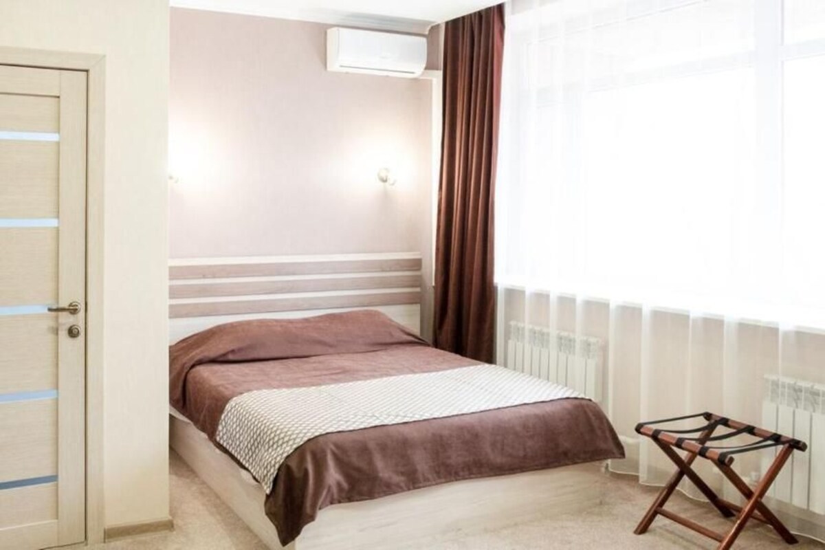 A bedroom with a bed, a radiator, a window with curtains, and a door with a light switch.