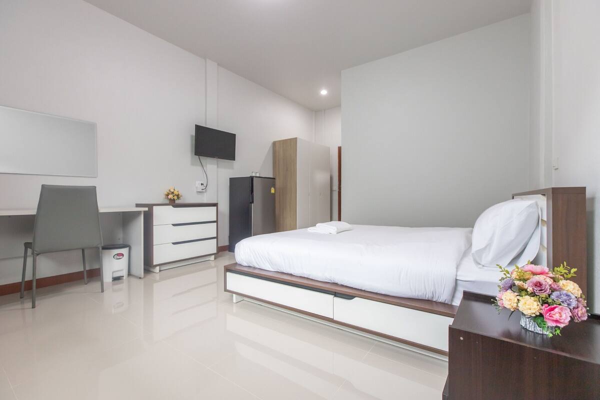 Ein modernes Hotelzimmer mit einem Bett, einem Schreibtisch, einem Stuhl, einem Fernseher, einem Kleiderschrank und einem kleinen Tisch mit Blumen.