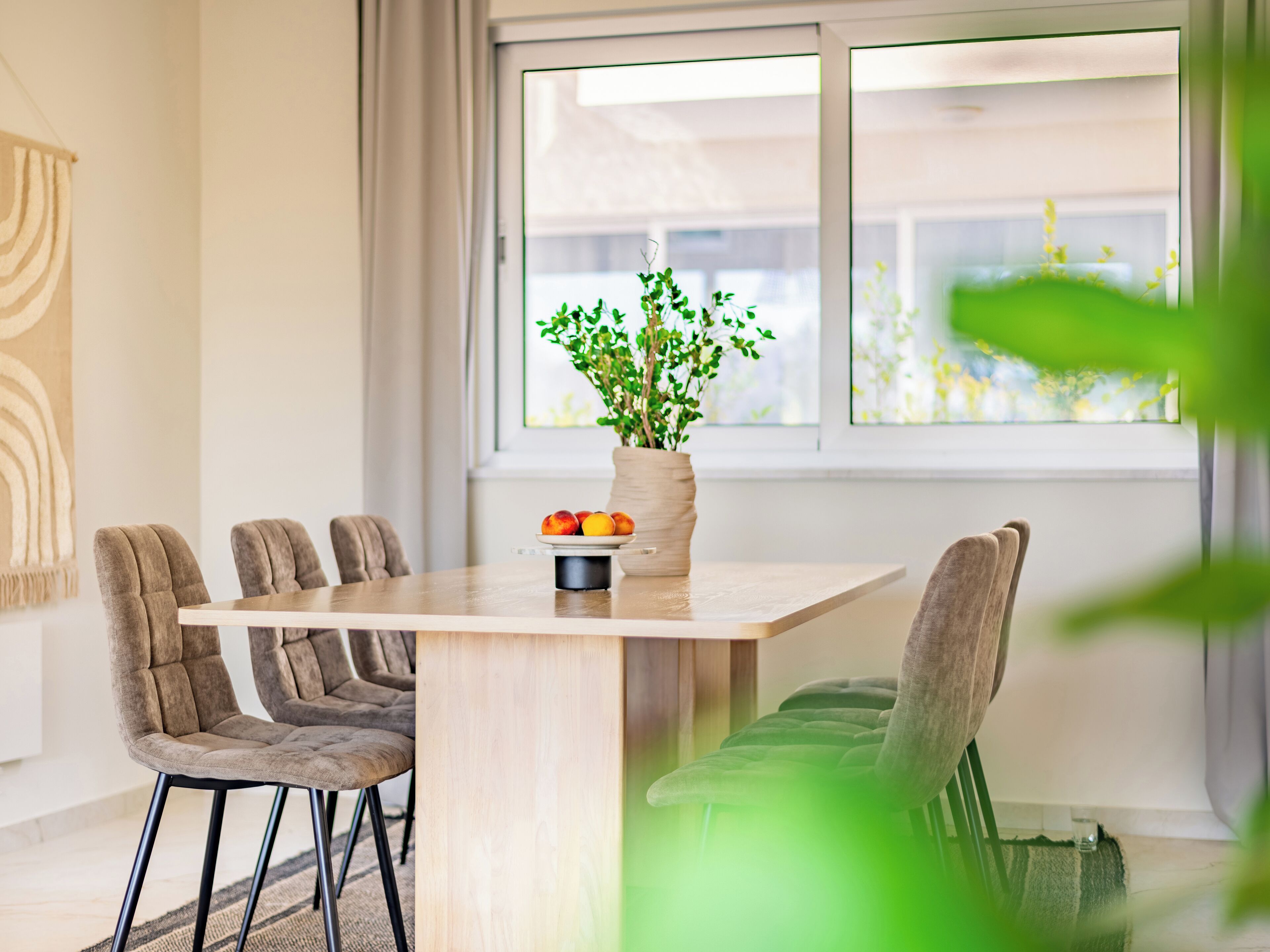 Un comedor con una mesa de madera, seis sillas tapizadas, un florero con plantas verdes y un plato de fruta.