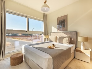 Relax in a spacious room with a large bed, featuring a quilted headboard and two pillows, and enjoy a view of the urban landscape from the large window.